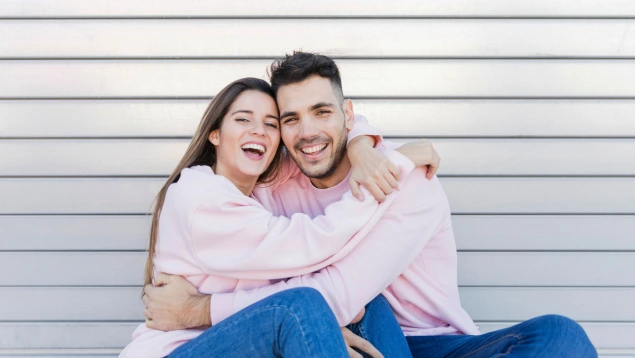 Pareja de hecho joven abrazándose y sonriendo con ropa a juego, sentados frente a una pared metálica.